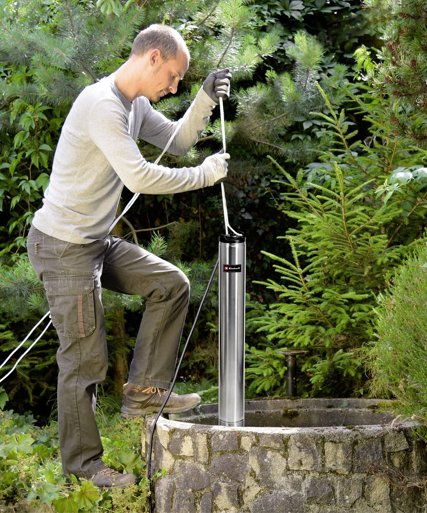 Ein Mann installiert eine Pumpe in einem Gartenbrunnen. Er trägt Arbeitshandschuhe und steht neben grünen Pflanzen.