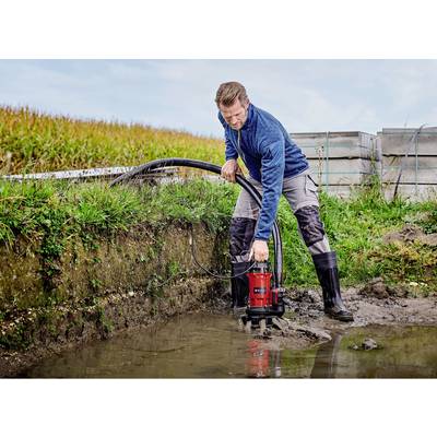 Ein Mann in Arbeitskleidung pumpt Wasser aus einem Graben mit einer kleinen, roten Wasserpumpe. Im Hintergrund ist ein Feld zu sehen.