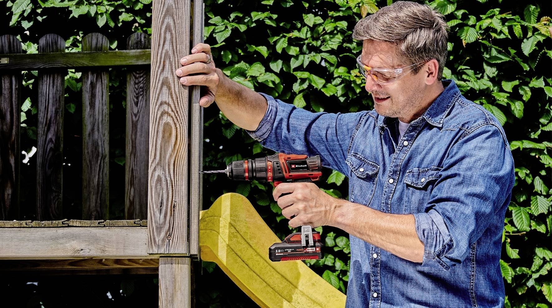 Eine Person benutzt eine Bohrmaschine, um Holz an einem Gartenzaun zu befestigen. Im Hintergrund sind grüne Büsche zu sehen.