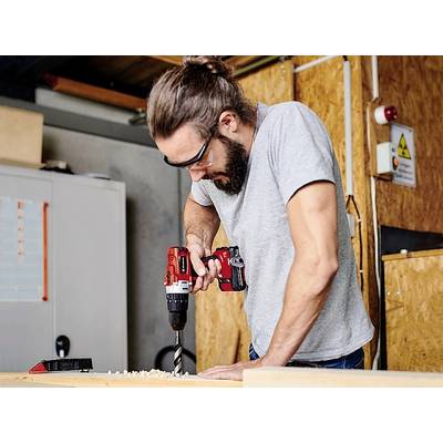 Ein Mann mit Schutzbrille arbeitet mit einem Akkuschrauber an einem Holzbrett in einer Werkstattumgebung.