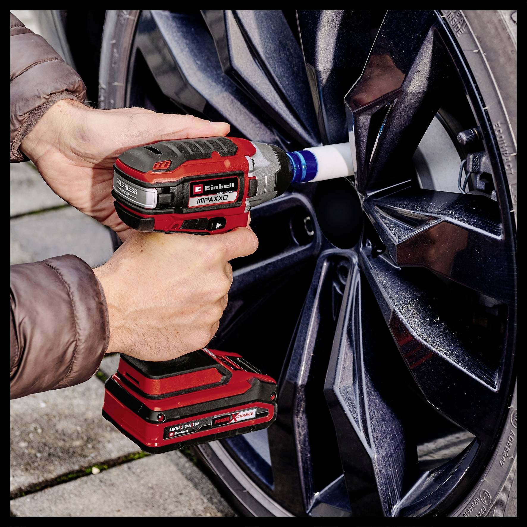 Eine Person mit brauner Jacke verwendet einen roten Akkuschlagschrauber an der Radmutter eines schwarzen Autos.