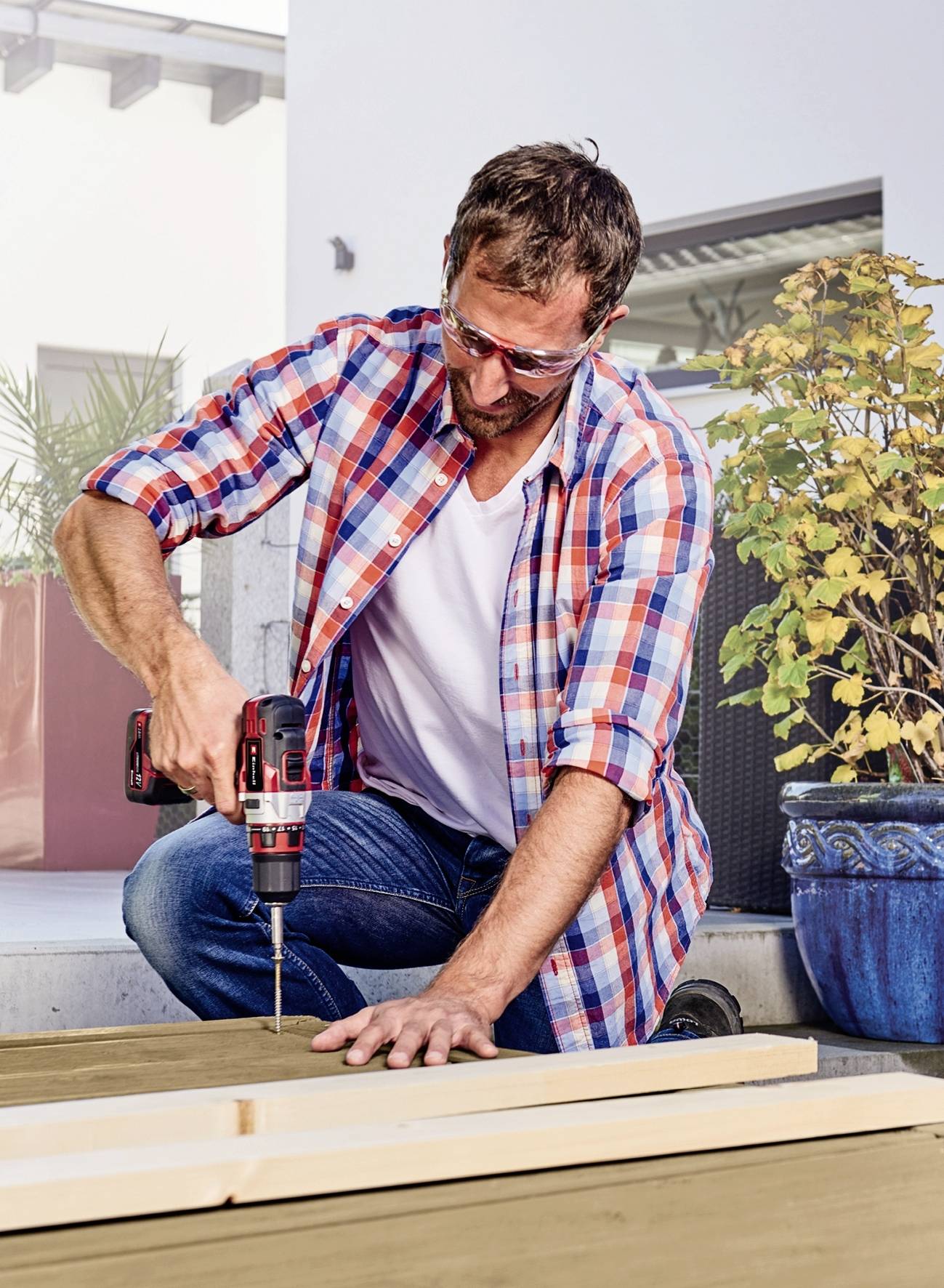 Ein Mann in kariertem Hemd bohrt mit einer elektrischen Bohrmaschine ein Loch in ein Holzbrett. Er trägt Schutzbrille. Im Freien.