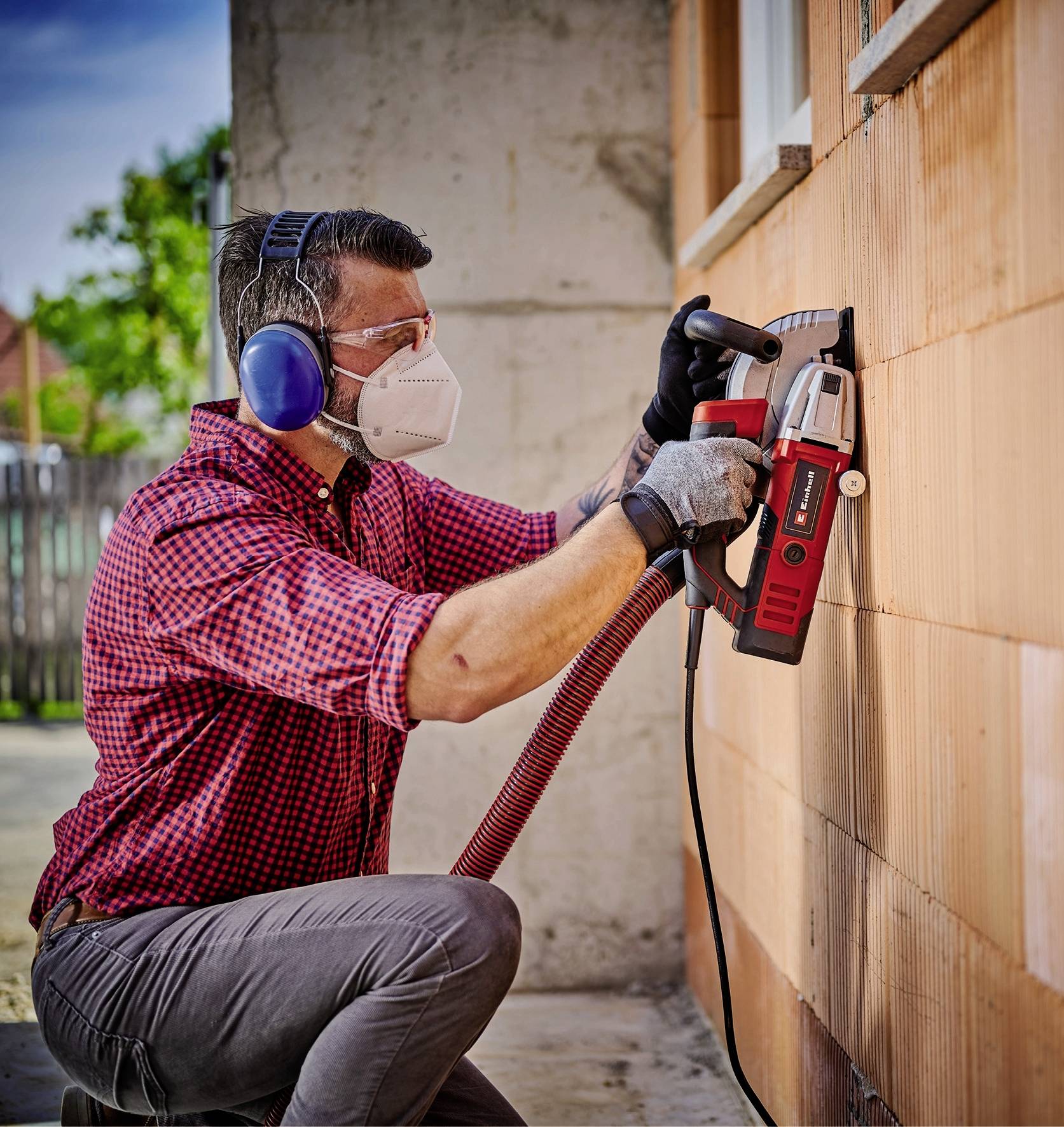 Ein Mann mit Gehörschutz und Maske bohrt mit einem Elektrowerkzeug ein Loch in eine Hauswand. Er trägt ein kariertes Hemd und Handschuhe.