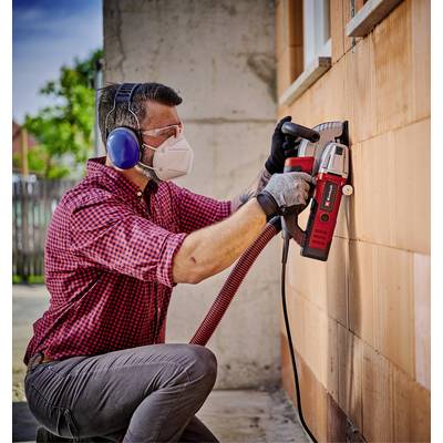 Ein Mann mit Gehörschutz und Maske bohrt mit einem Elektrowerkzeug ein Loch in eine Hauswand. Er trägt ein kariertes Hemd und Handschuhe.