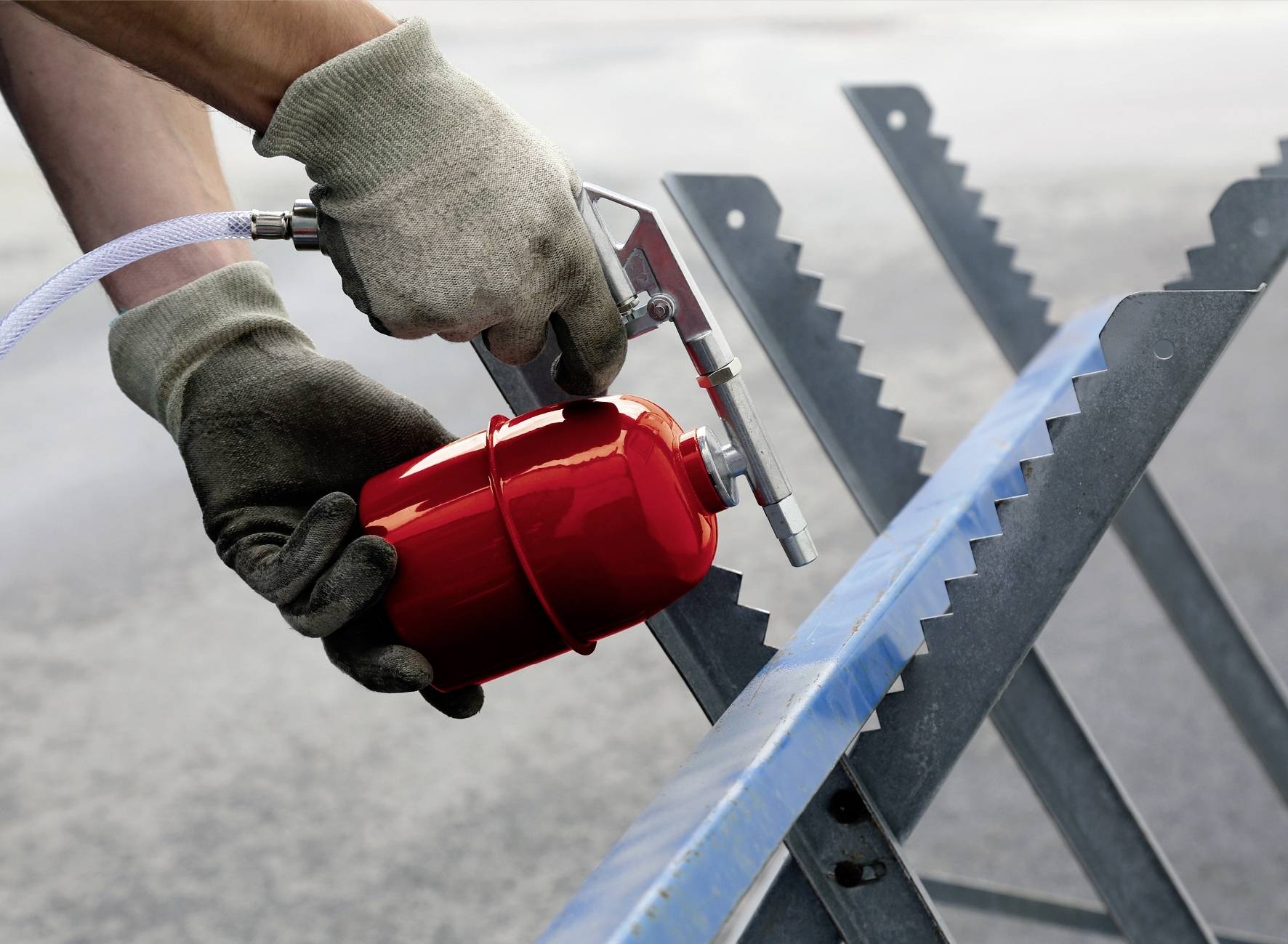 Eine Person trägt Handschuhe und sprüht ein rotes Lackierwerkzeug auf eine Metalloberfläche, vermutlich zur Beschichtung oder zum Schutz.