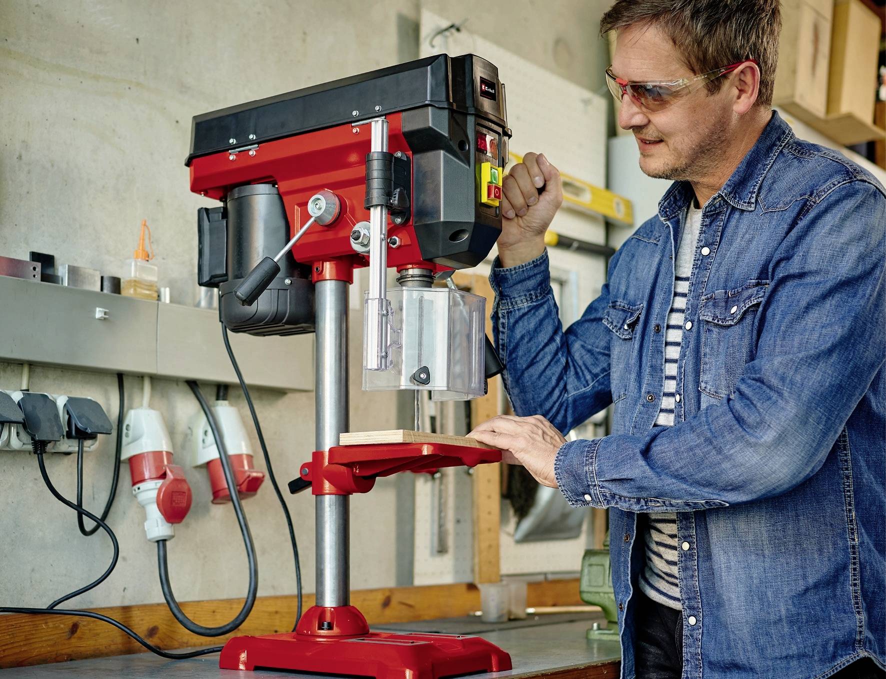Ein Mann in Schutzbrille bearbeitet Holz mit einer Tischbohrmaschine in einer Werkstatt.