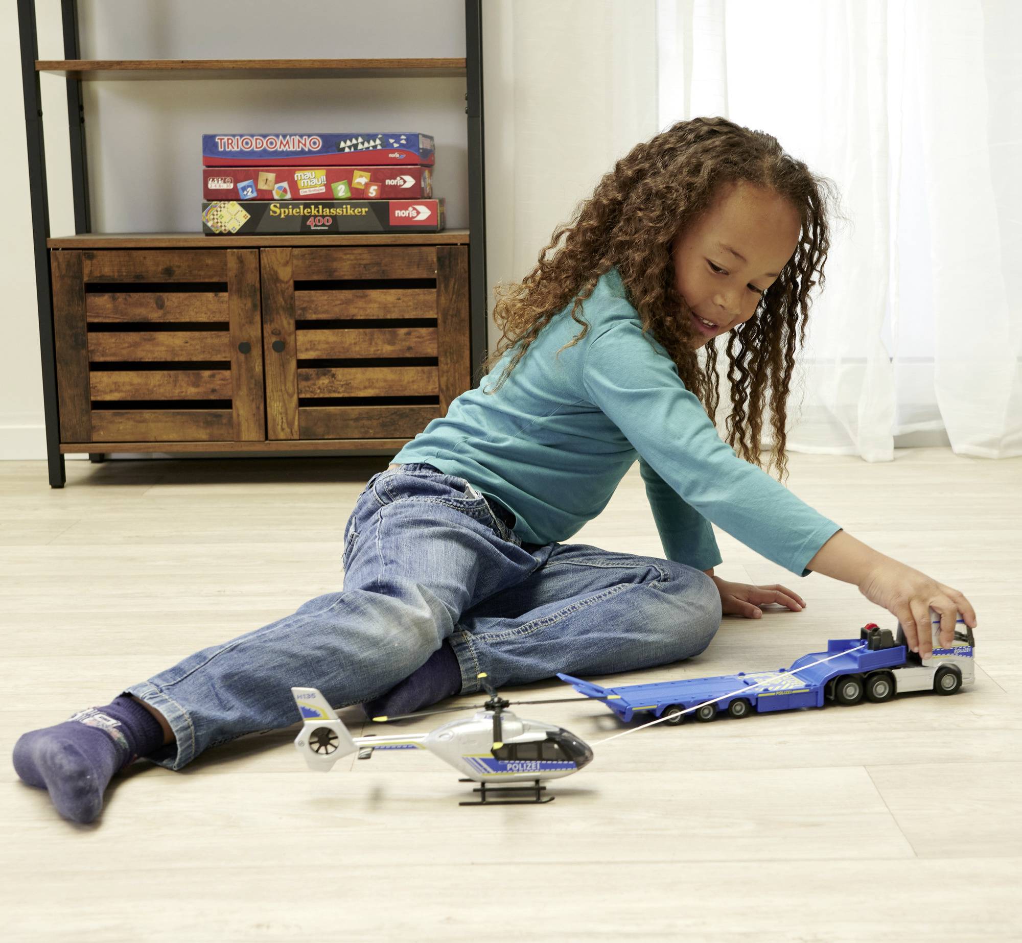 Ein Kind spielt auf dem Boden mit einem Spielzeug-LKW und einem Hubschrauber. Im Hintergrund sind Brettspiele auf einem Regal.