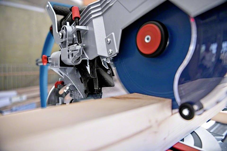 Eine Kreissäge schneidet durch ein Holzbrett. Die rotierende blaue Klinge ist teilweise sichtbar, Metall- und Kunststoffteile umgeben sie.