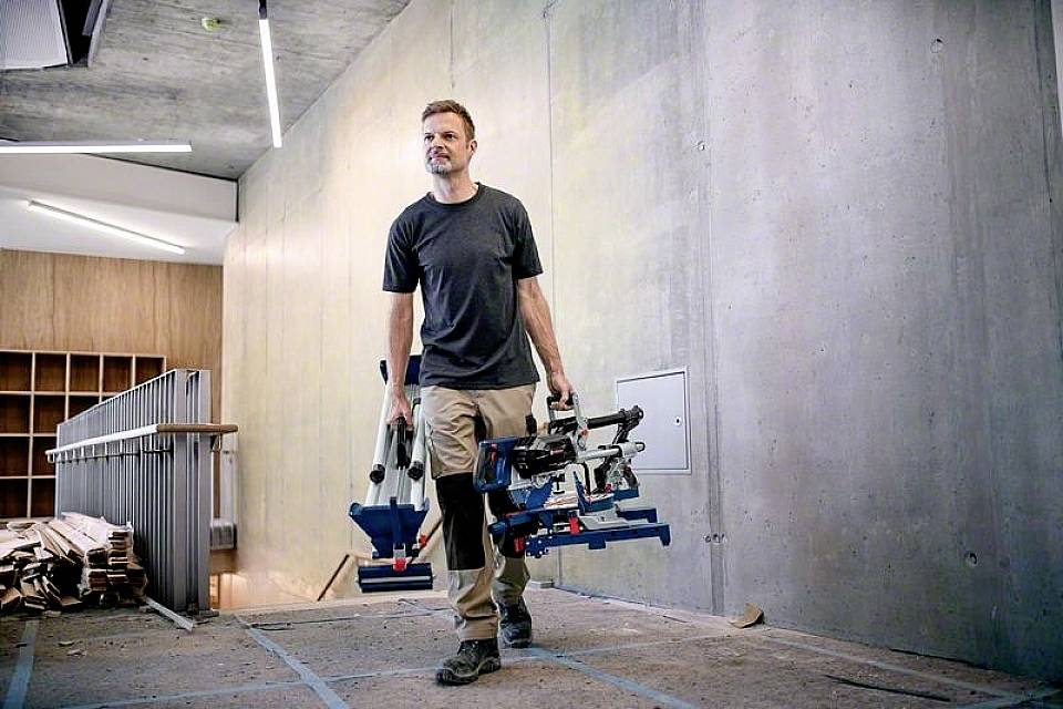 Eine Person trägt Werkzeuge in einem Gebäude, das im Bau oder in der Renovierung ist. Im Hintergrund sind Betonwände und Holzregale sichtbar.