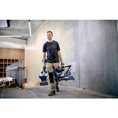 Eine Person trägt Werkzeuge in einem Gebäude, das im Bau oder in der Renovierung ist. Im Hintergrund sind Betonwände und Holzregale sichtbar.
