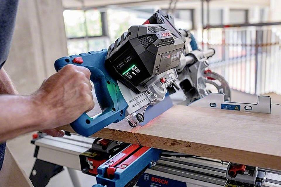 Eine Person benutzt eine Bosch-Handkreissäge, um ein Holzbrett präzise zuzuschneiden. Die Säge zeigt digitale Informationen an.