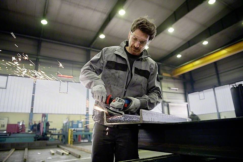 Ein Arbeiter in Schutzkleidung schleift mit einem Winkelschleifer Metall in einer Fabrikhalle. Funken sprühen vom Metall ab.