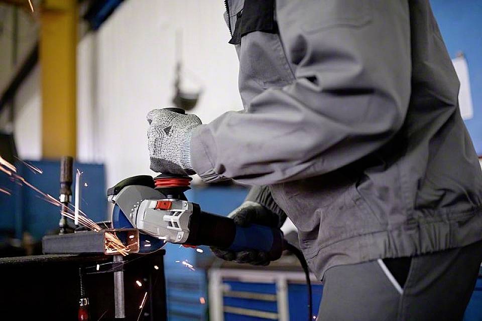 Eine Person in Schutzkleidung schleift Metall mit einem Winkelschleifer, Funken fliegen. Sie arbeitet in einer Werkstatt.