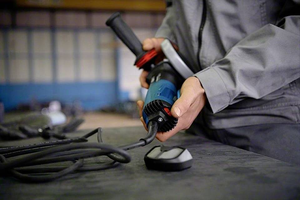 Eine Person in Arbeitskleidung hält eine blaue Elektrowerkzeug-Maschine mit Kabel, wahrscheinlich in einer Fabrikumgebung.