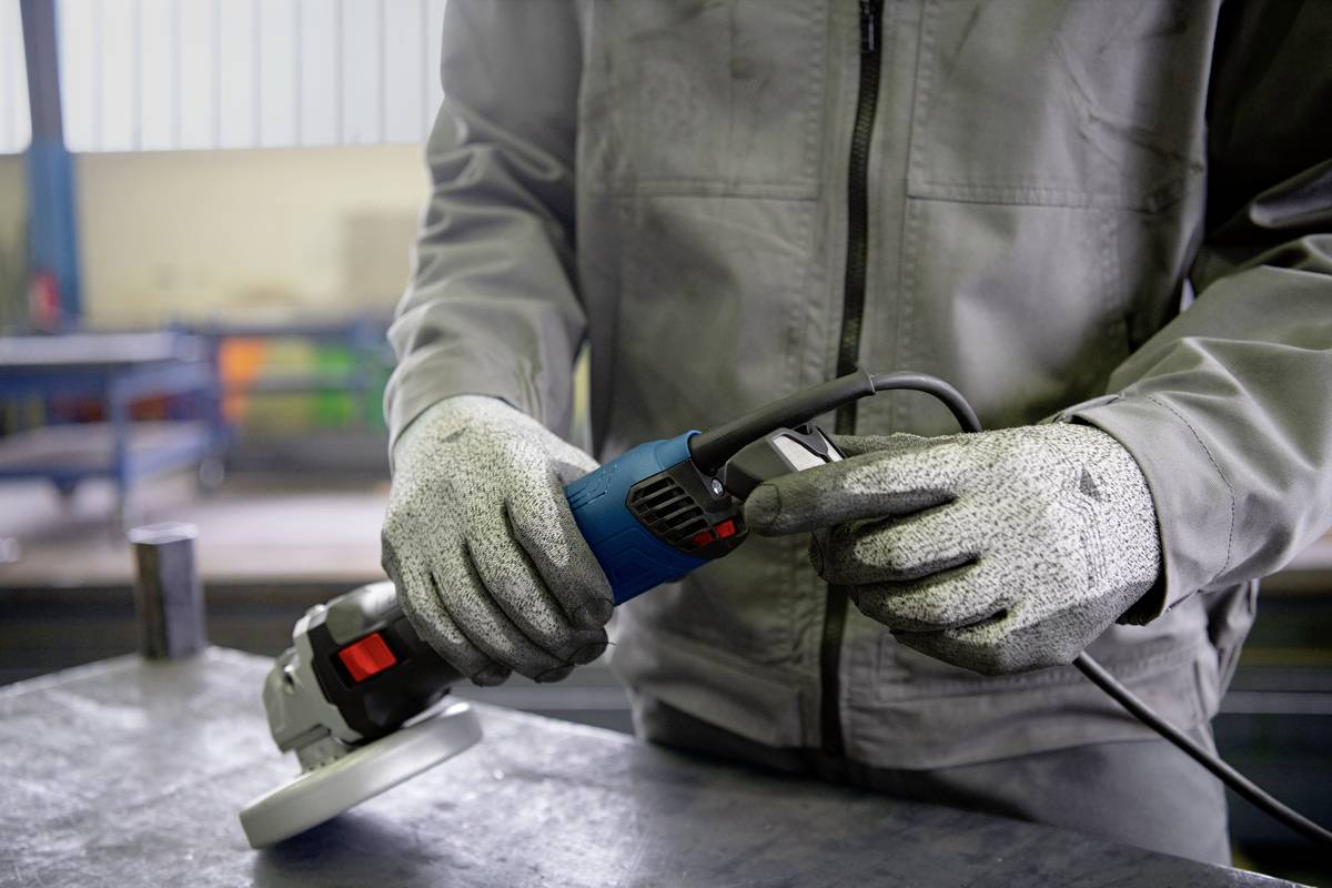 Eine Person in Arbeitskleidung und Handschuhen hält und bedient ein elektrisches Werkzeug zur Metallbearbeitung in einer Werkstattumgebung.