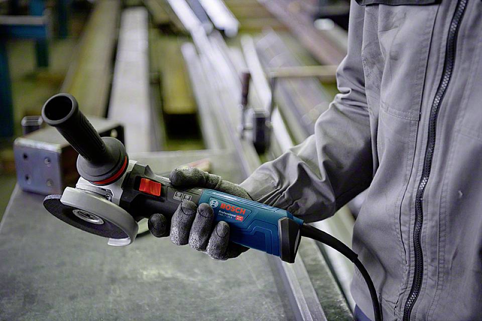 Eine Person hält einen Bosch-Winkelschleifer in der Hand. Im Hintergrund sind Metallteile in einer Werkstatt sichtbar.