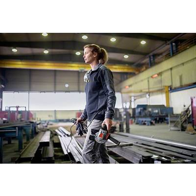 Eine Frau in Arbeitskleidung steht in einer Fabrikhalle und hält ein Elektrowerkzeug. Die Halle ist groß und industriell.