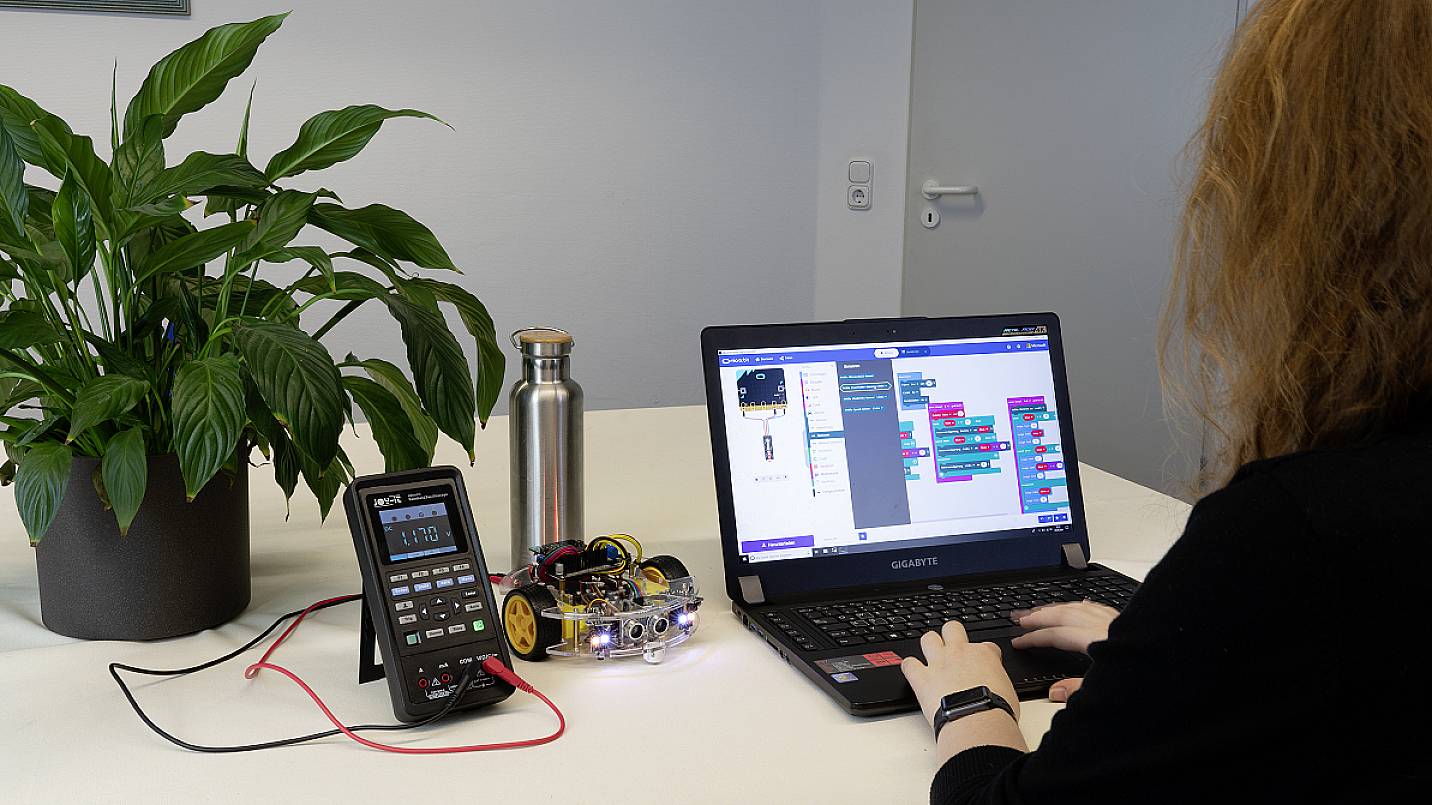 Eine Person programmiert einen Roboter mithilfe eines Laptops. Ein Oszilloskop und eine Pflanze sind auf dem Tisch sichtbar.