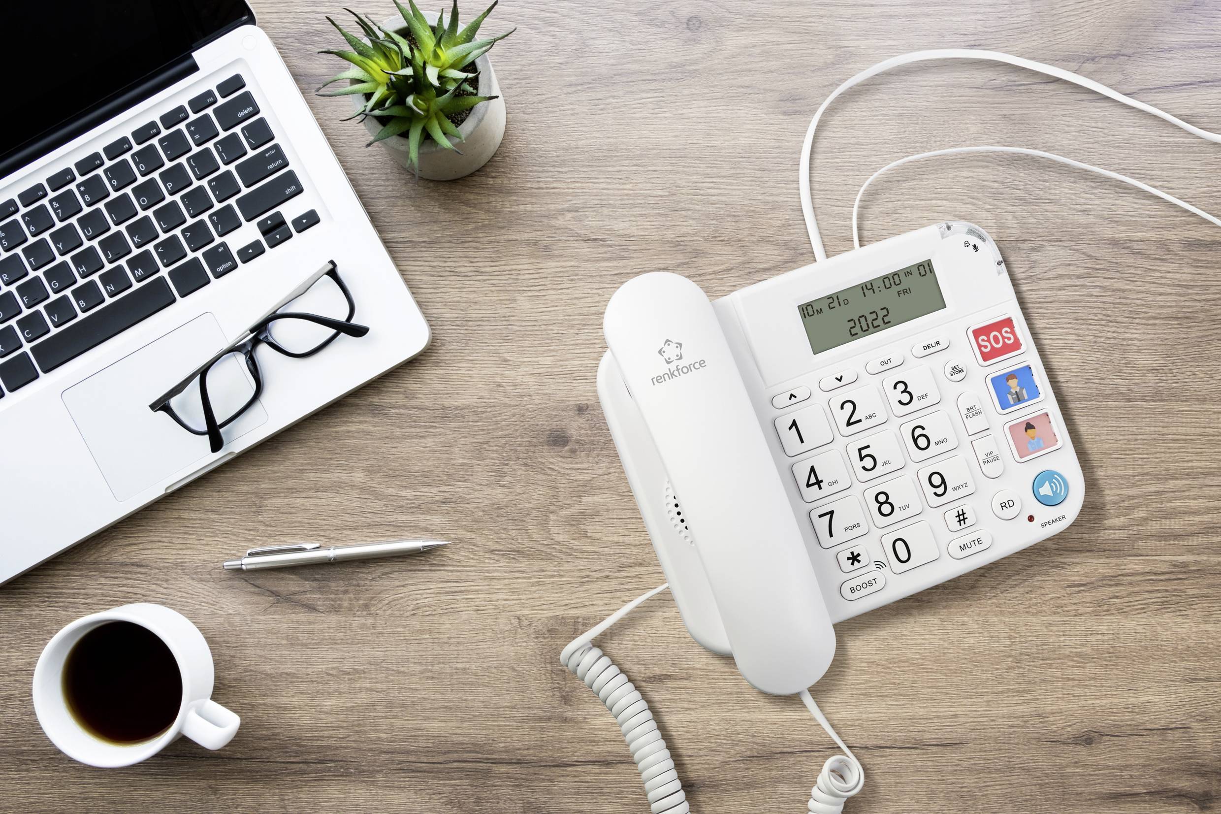 Auf einem Schreibtisch befinden sich ein Laptop, eine Brille, ein Stift, ein Telefon mit großen Tasten, eine Tasse Kaffee und eine Pflanze.