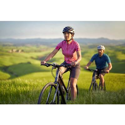 Zwei Personen fahren auf Fahrrädern durch eine grüne, hügelige Landschaft. Im Vordergrund eine Frau mit rotem Trikot.