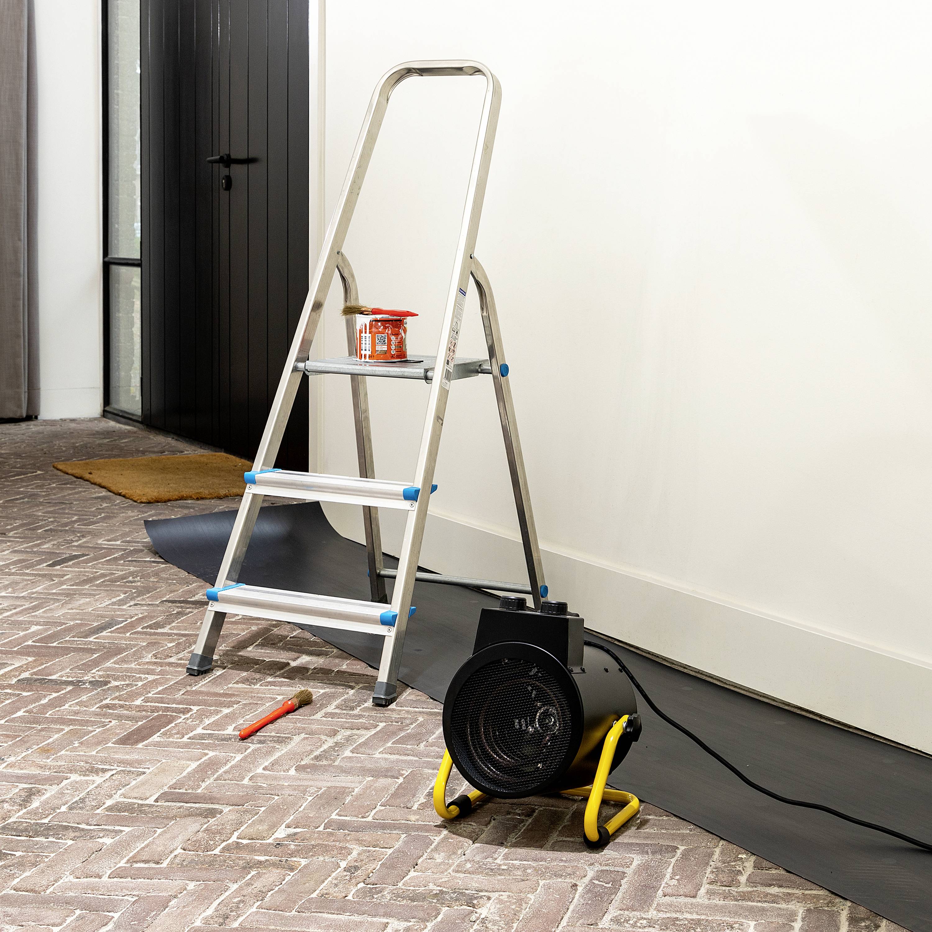 Eine Leiter steht auf einem gefliesten Boden neben einer Wand, die renoviert wird. Auf der Leiter steht ein Farbeimer mit einem Pinsel.