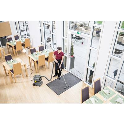 Eine Person saugt eine Matte am Eingang eines hellen, leeren Restaurants. Die Tische sind mit Tischsets und Servietten gedeckt. Große Fenster bieten Ausblick nach draußen.
