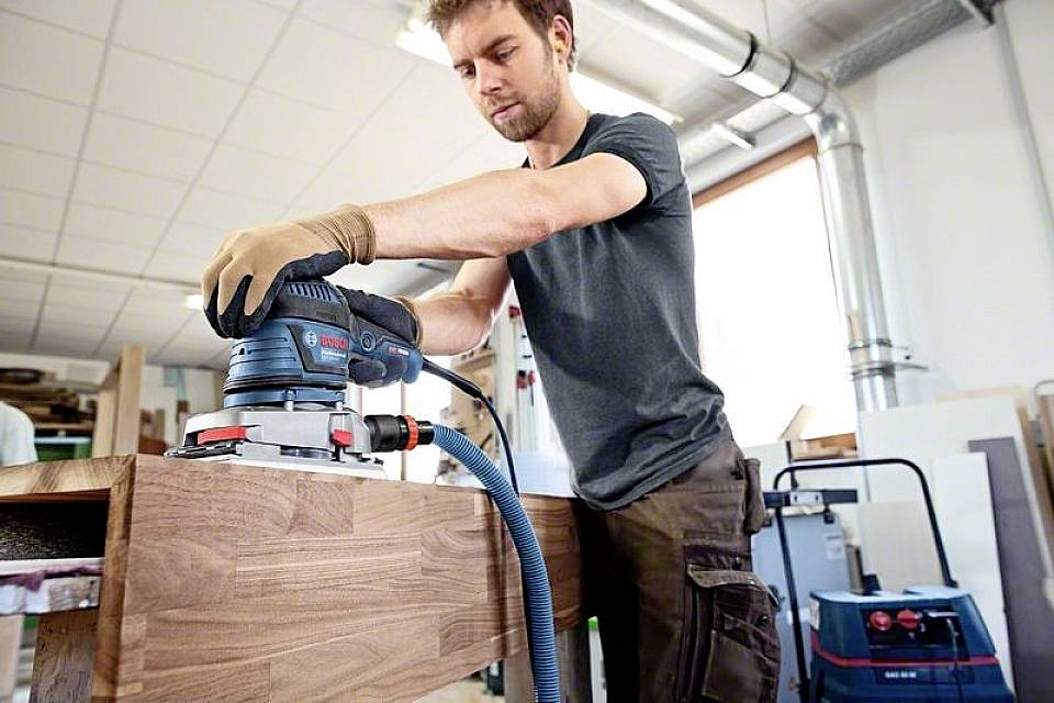 Ein Mann schleift eine Holzkante in einer Werkstatt mit einer elektrischen Schleifmaschine, umgeben von Werkzeugen und Geräten.