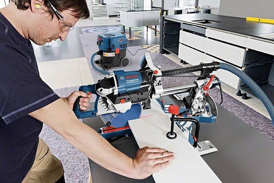 Eine Person bedient eine Sägemaschine in einer Werkstatt. Die Maschine schneidet ein weißes Brett. Im Hintergrund steht ein Staubsauger.