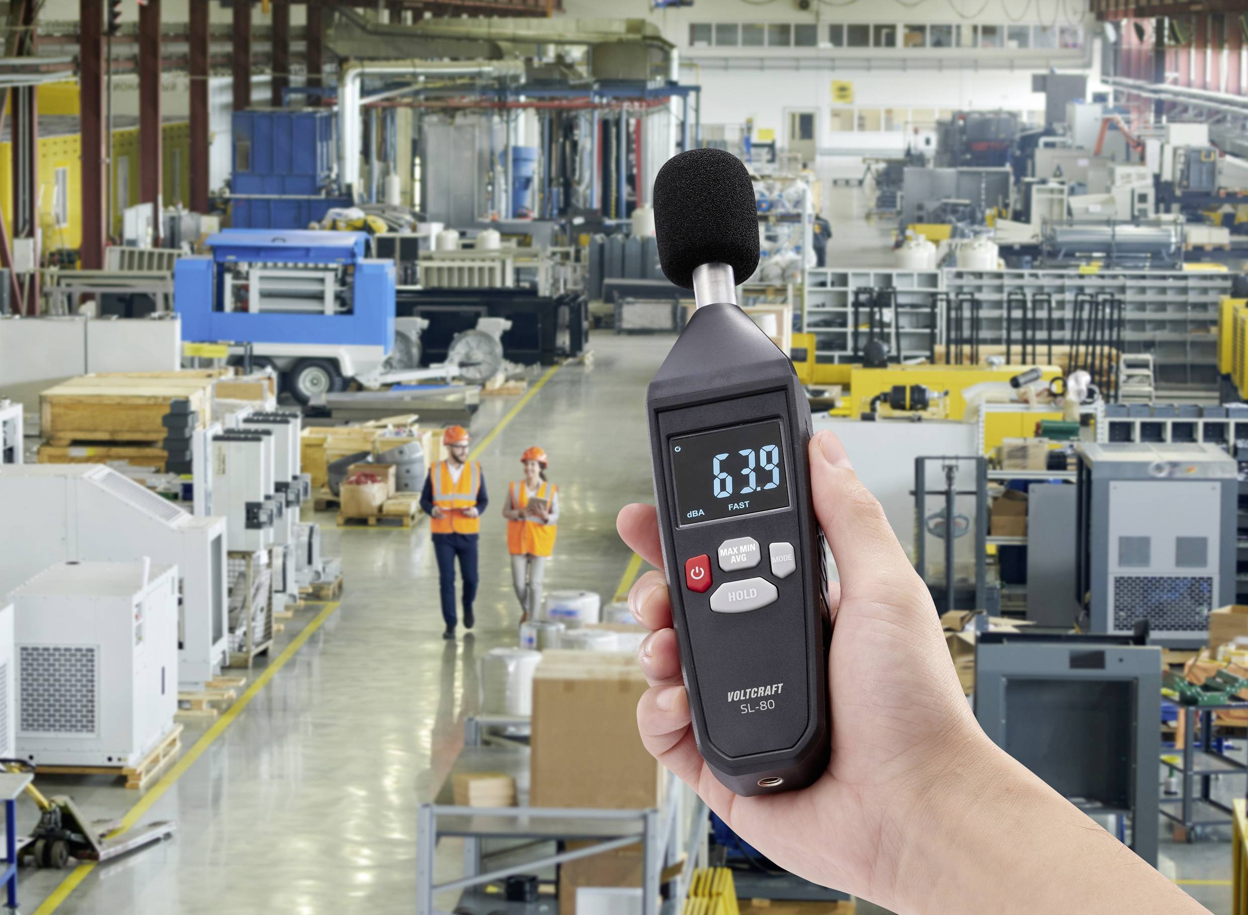 Eine Person hält ein Dezibelmessgerät, das 63,9 dB anzeigt, in einer großen Fabrikumgebung mit Maschinen und Mitarbeitern im Hintergrund.