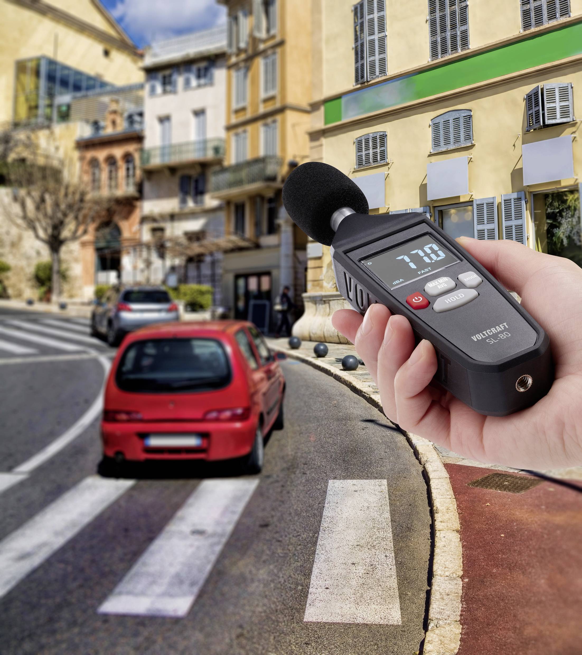 Eine Person misst die Lautstärke mit einem Schallpegelmessgerät neben einer belebten Straße mit vorbeifahrendem roten Auto.