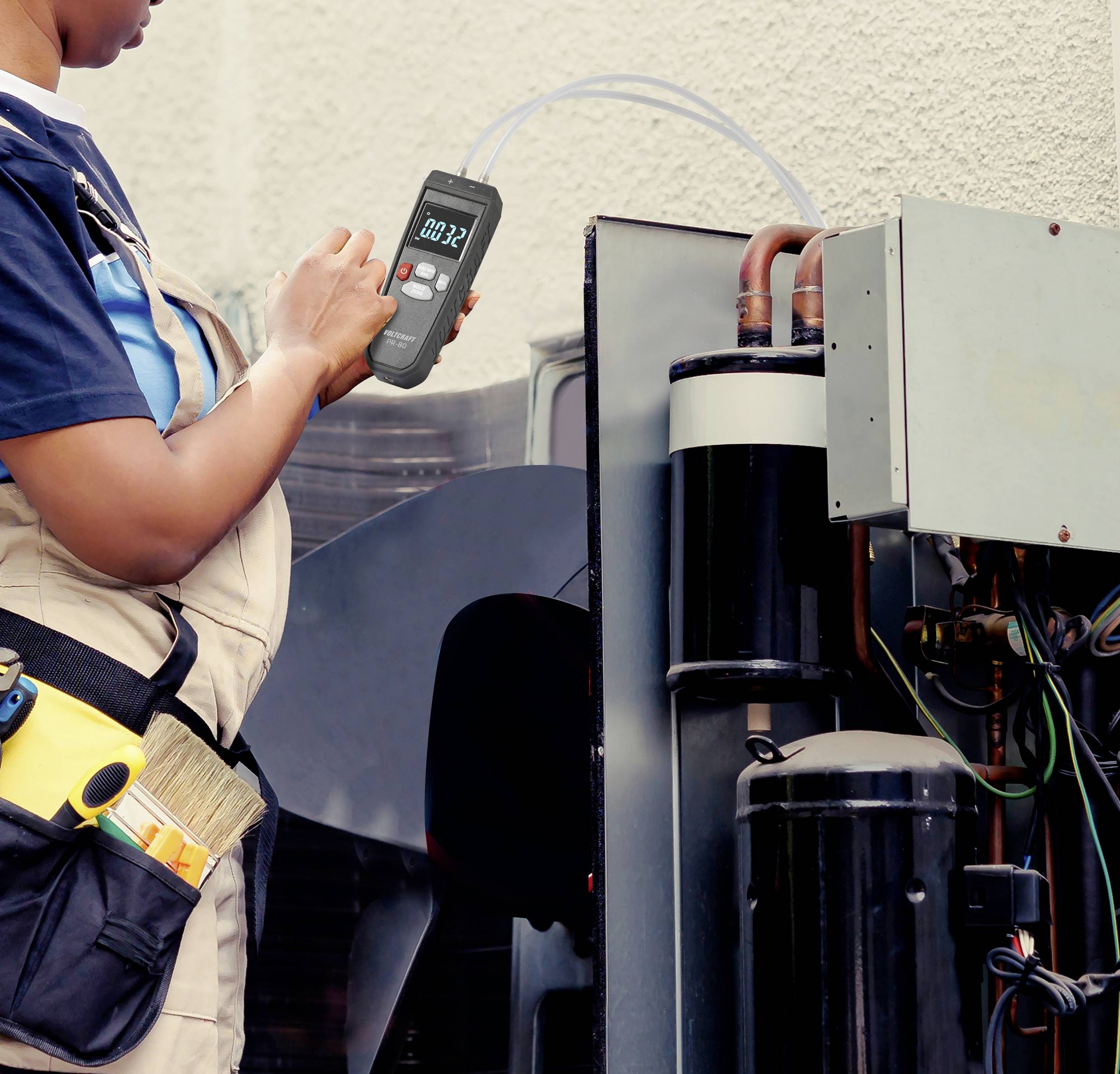 Eine Person überprüft den Luftdruck an einer Klimaanlage mit einem Messgerät. Sie trägt eine Werkzeugtasche.