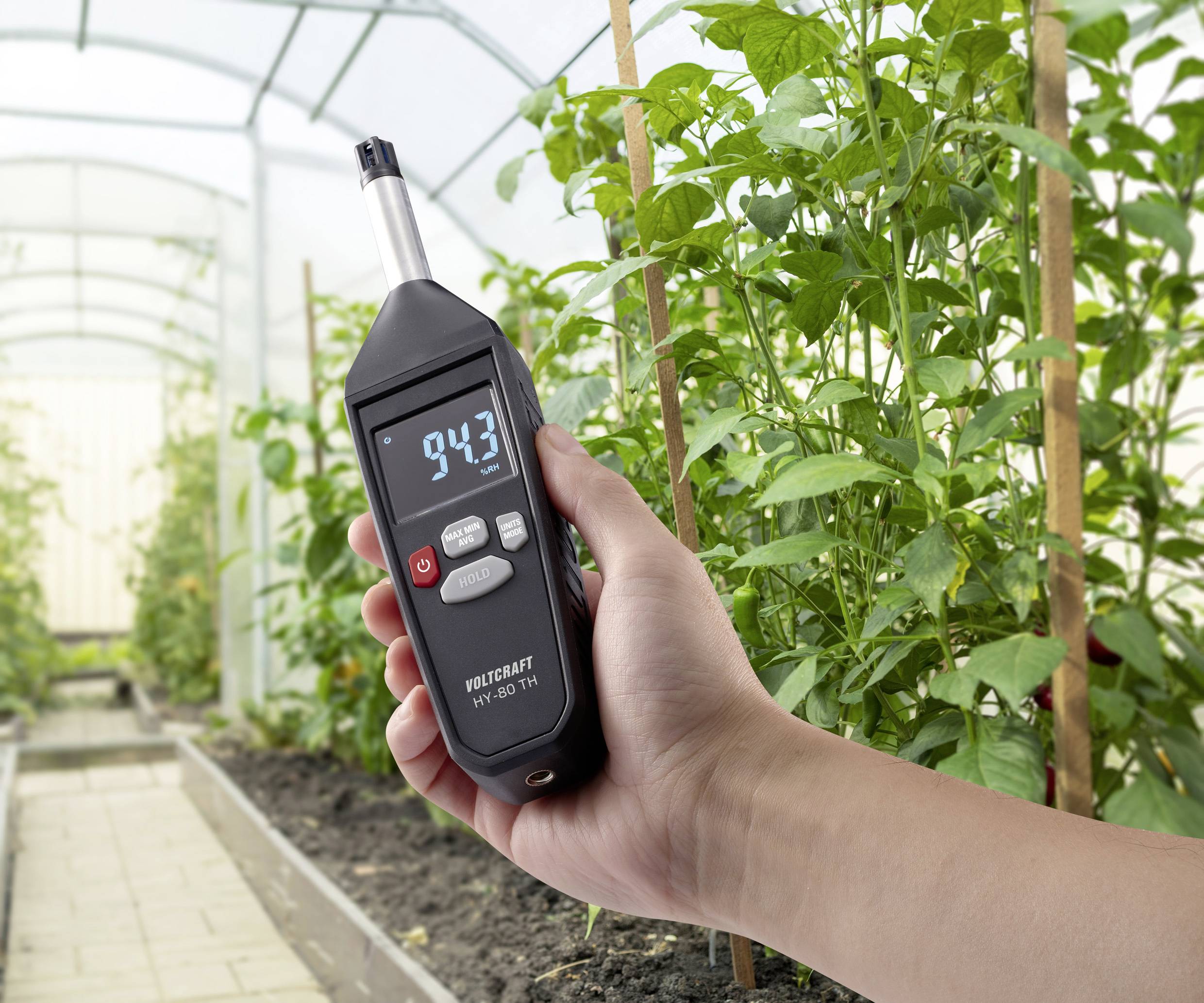 Eine Hand hält ein digitales Messgerät im Gewächshaus, das eine Anzeige von '94,3' zeigt. Pflanzen wachsen im Hintergrund.