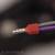 Ein rot ummanteltes Kabelende mit silberner, wellenförmiger Metallspitze, vor unscharfem Hintergrund.