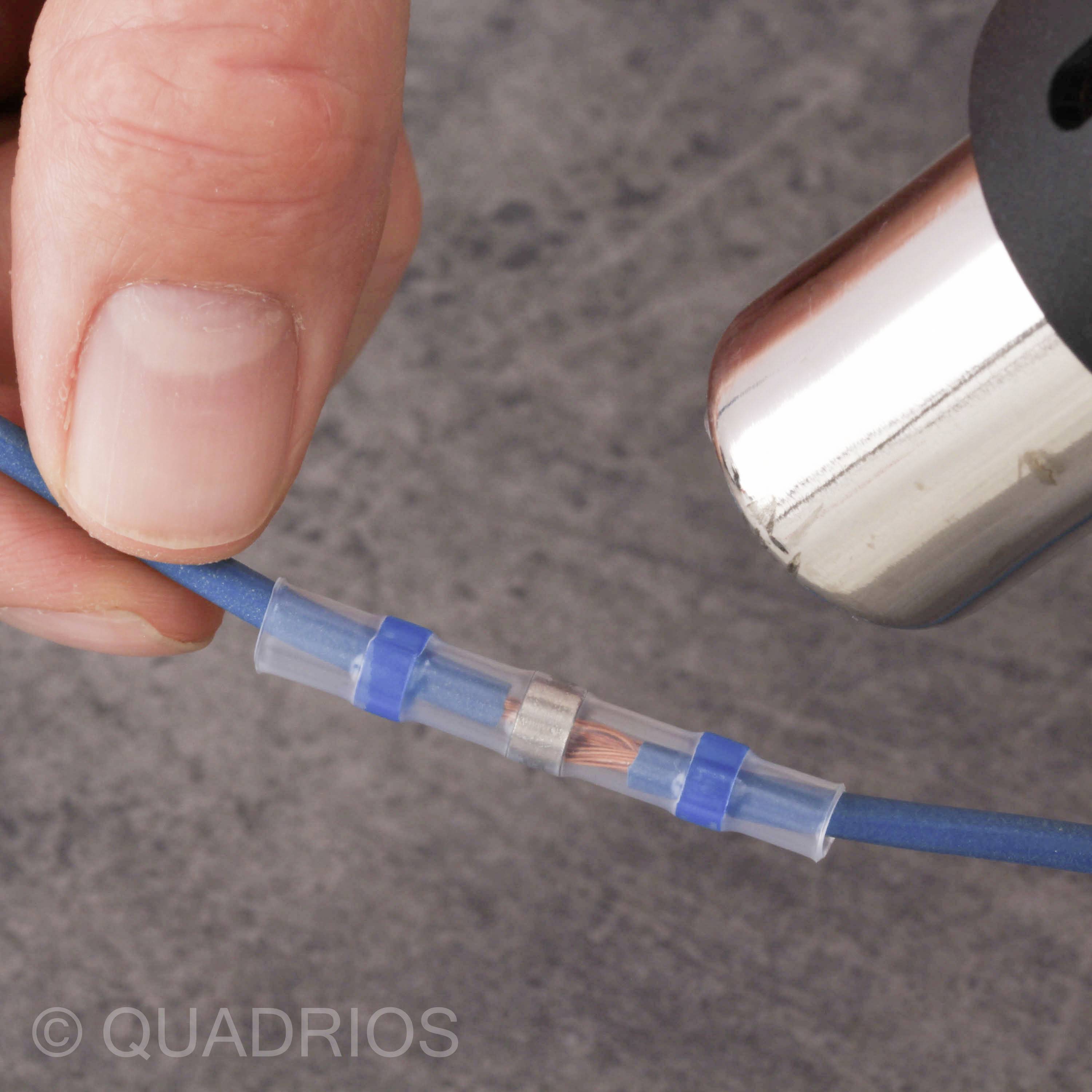Eine Hand hält ein blaues Kabel. Das Kabel wird mit einer Klemmverbindung verbunden. Ein Föhn erhitzt die Verbindung.