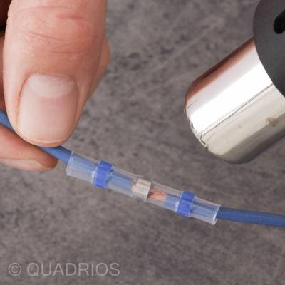 Eine Hand hält ein blaues Kabel. Das Kabel wird mit einer Klemmverbindung verbunden. Ein Föhn erhitzt die Verbindung.