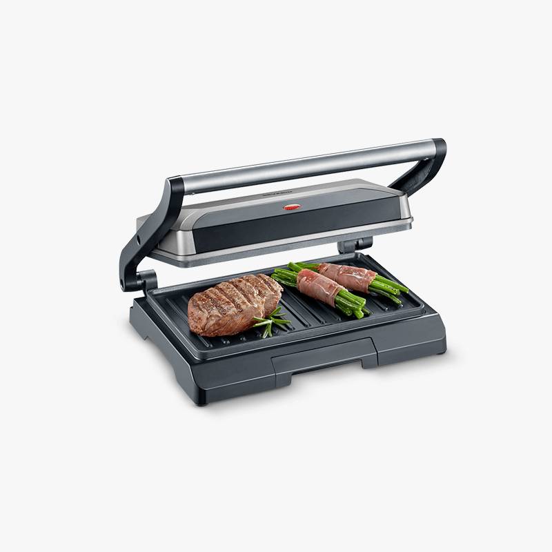 Ein elektrischer Kontaktgrill mit geöffnetem Deckel grillt zwei Steaks und grüne Spargelstangen umwickelt mit Speck.