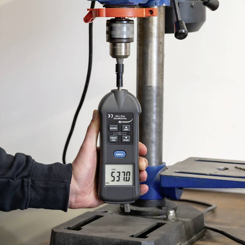 Eine Person hält ein Digitalmessgerät unter einer Bohrmaschine, um die Drehzahl zu messen.