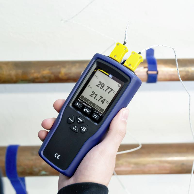 Eine Hand hält ein digitales Thermometer, das zwei Messungen anzeigt: 21,74°C und 25,94°C. Hintergrund zeigt Rohrleitungen.
