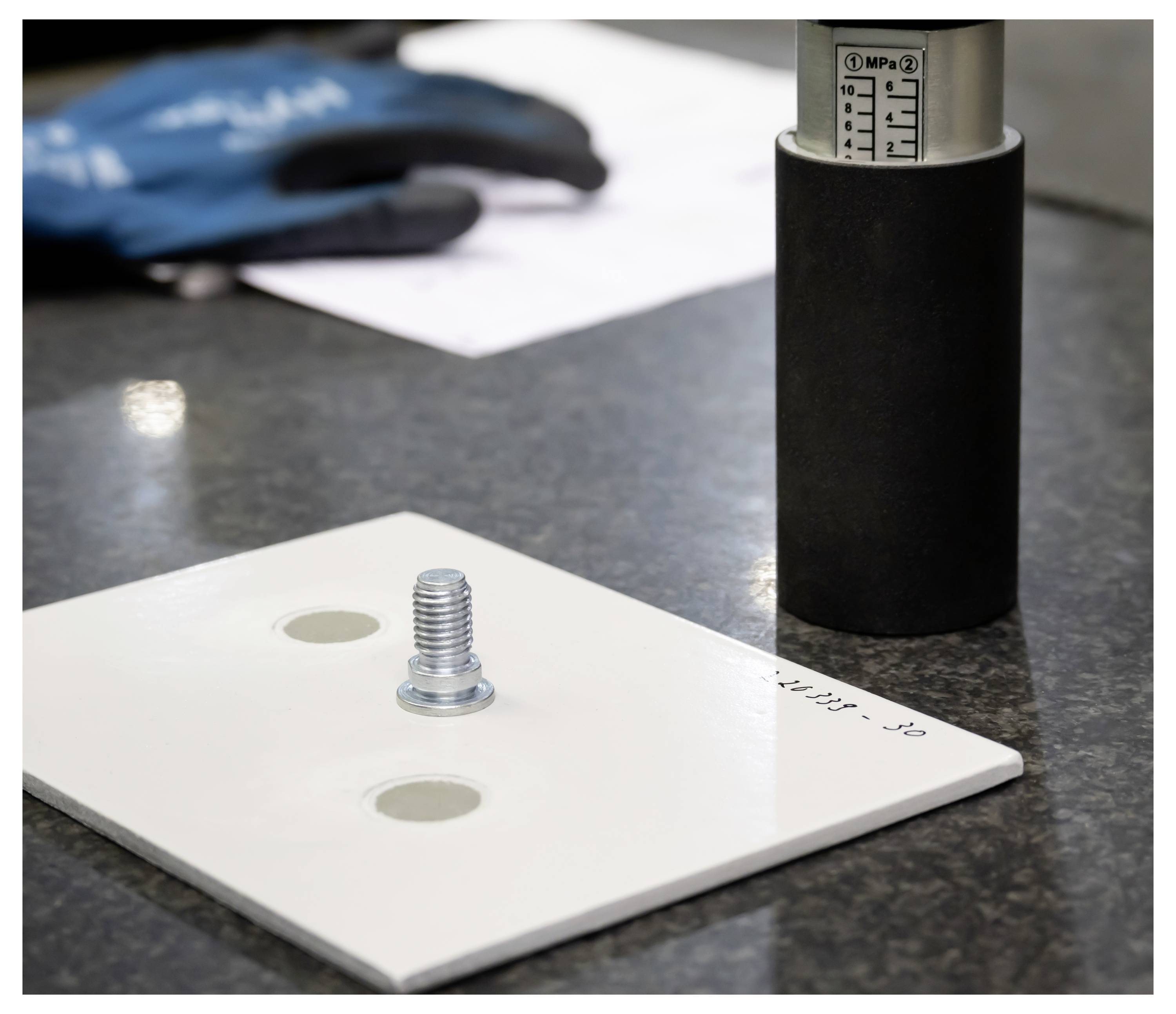 Weißes Testmuster mit einem Gewinde-Metallstift, der neben einem Druckmanometer in einer Labor- oder Inspektionseinrichtung verwendet wird.