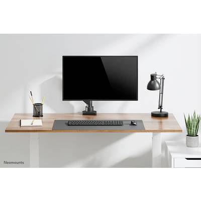 Ein aufgeräumter Schreibtisch mit Monitor, Tastatur, Maus, Schreibtischlampe, Stifthalter, Notizblock und Pflanze auf hellem Holzuntergrund.