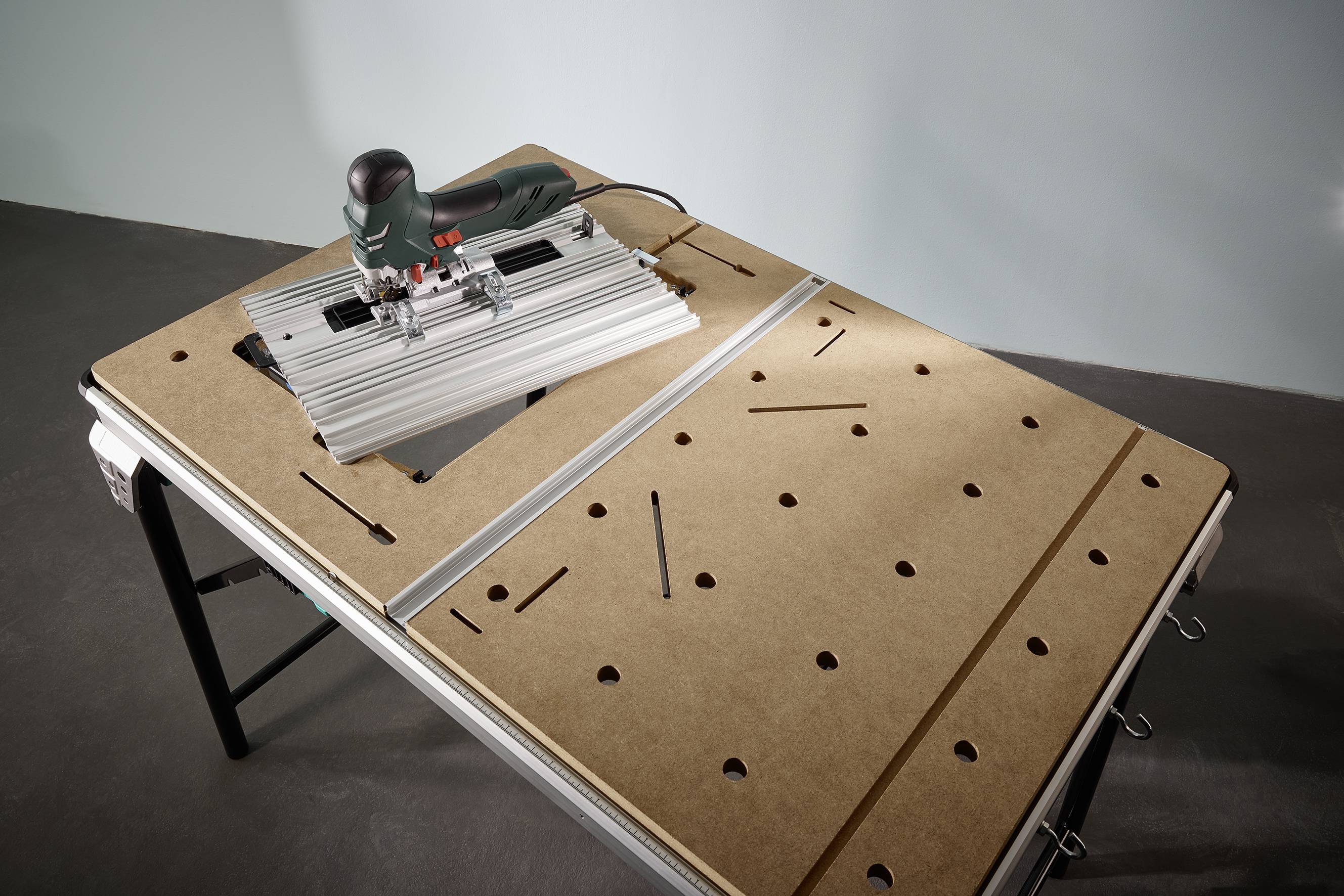Eine elektrische Stichsäge auf einem multifunktionalen Werkstatttisch mit Holzplatte. Der Tisch hat Löcher und Schienen zur flexiblen Nutzung.