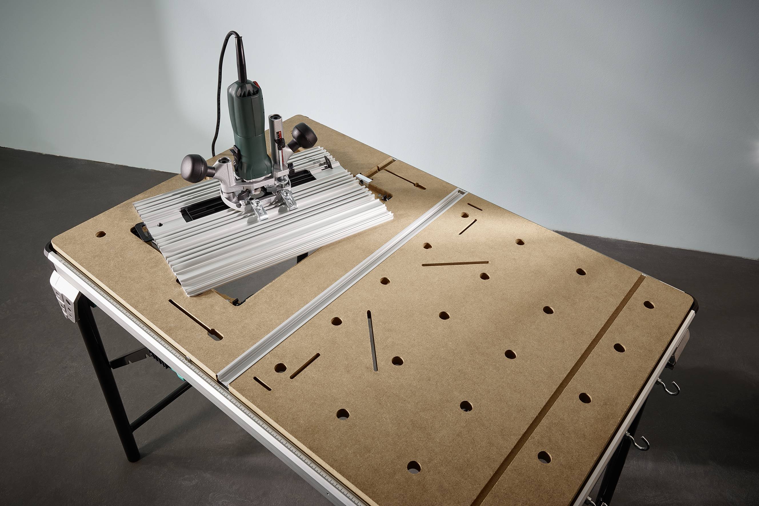 'Oberfräse auf Werkbank in Werkstatt. Gerät mit grünem Gehäuse befestigt auf einer Klemmschiene, umgeben von montierbaren Platten.'