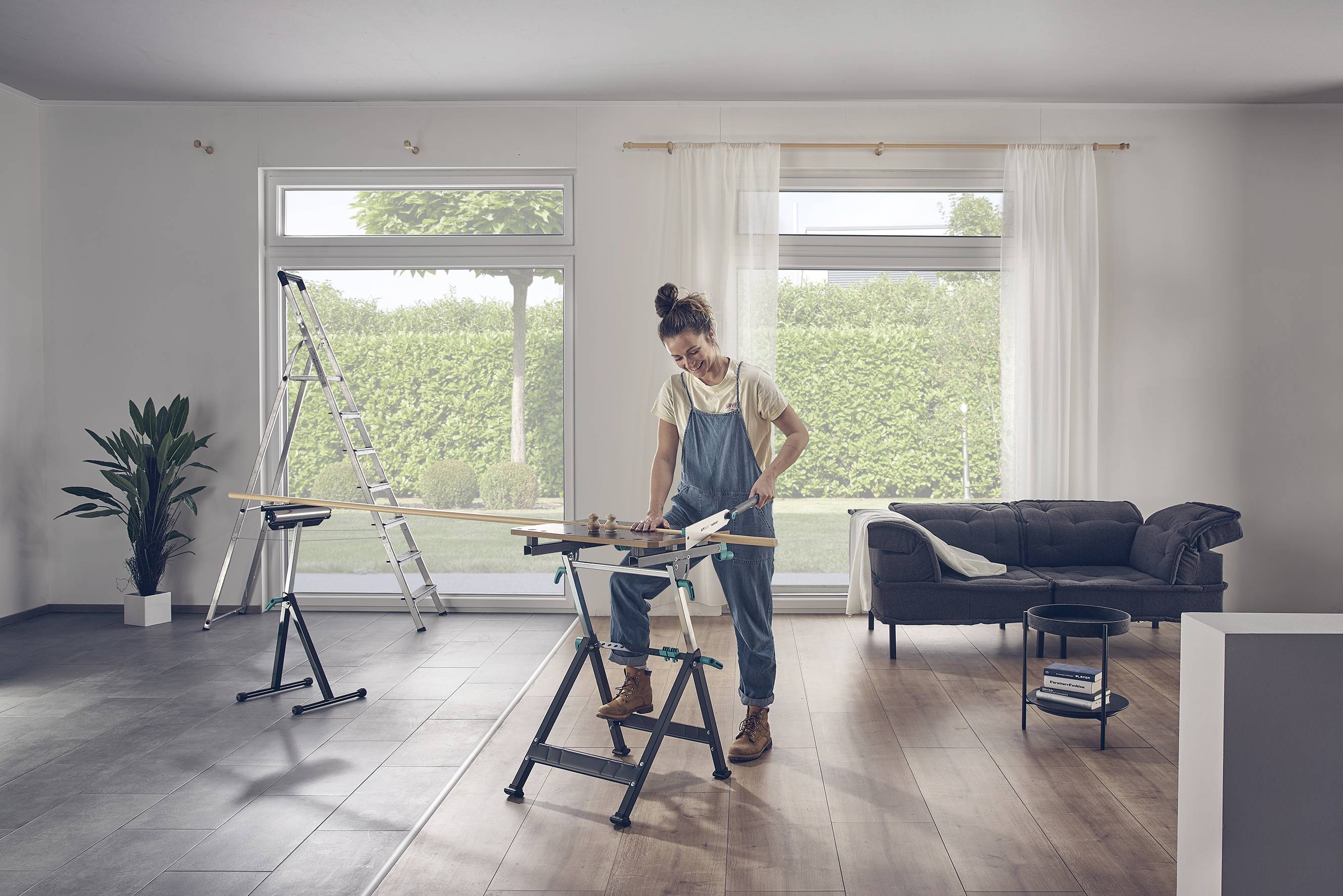 Eine Frau in Arbeitskleidung arbeitet mit Werkzeug an einem Holzbalken in einem Wohnzimmer. Eine Leiter steht neben ihr, ein Sofa im Hintergrund.