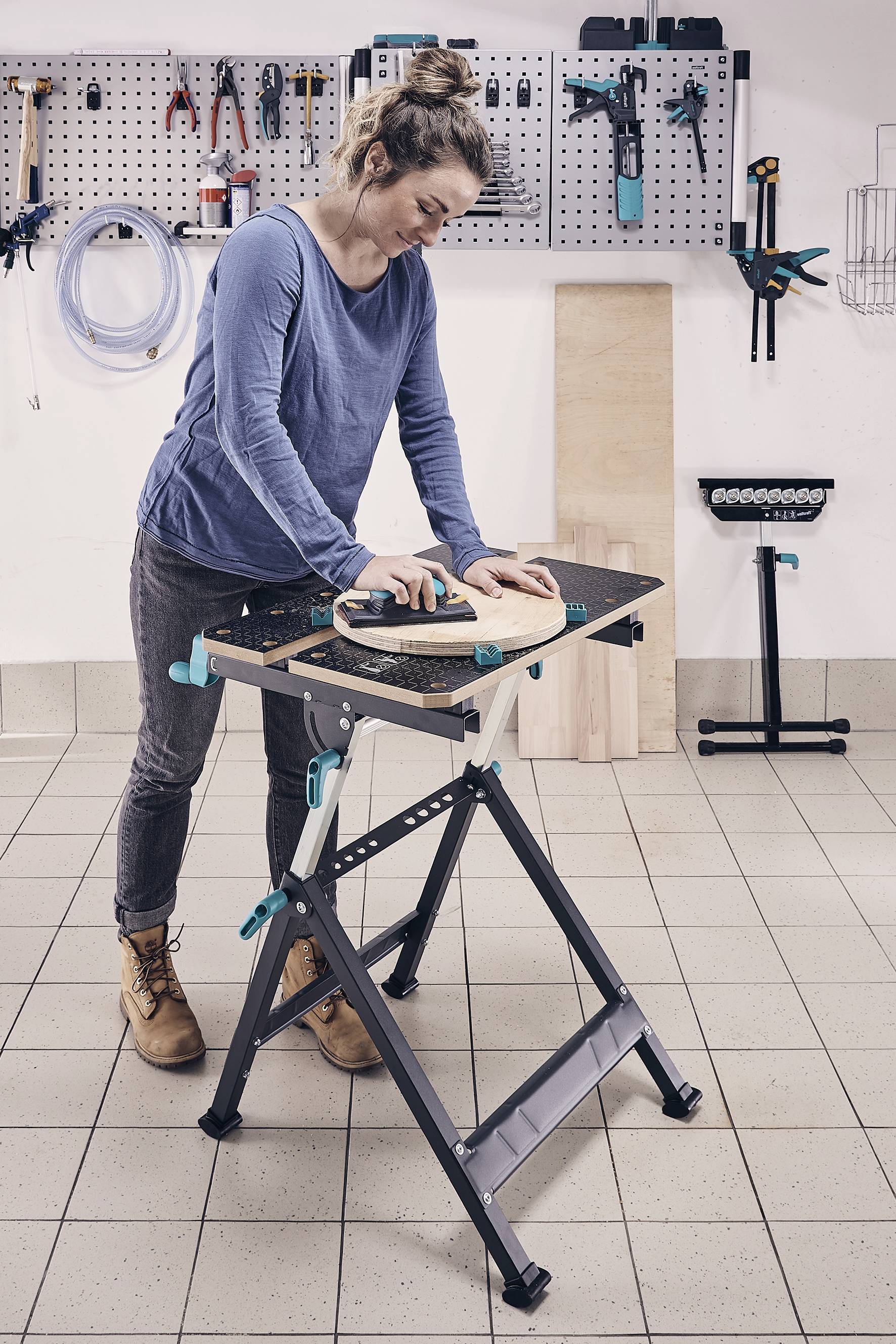 Eine Frau arbeitet in einer Werkstatt an einer Werkbank und schleift ein rundes Holzstück. Im Hintergrund sind Werkzeuge an der Wand.