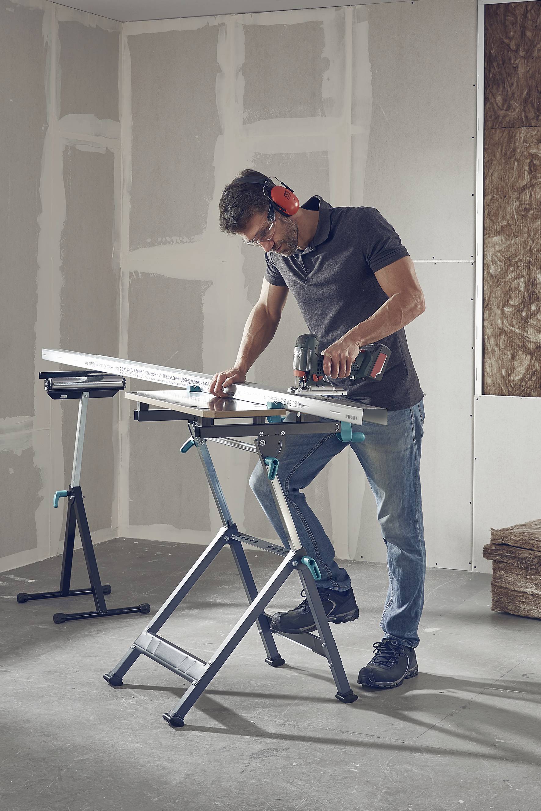 Ein Mann arbeitet mit Gehörschutz und Schutzbrille in einem teilausgebauten Raum. Er benutzt eine elektrische Säge, um ein Brett zu schneiden.