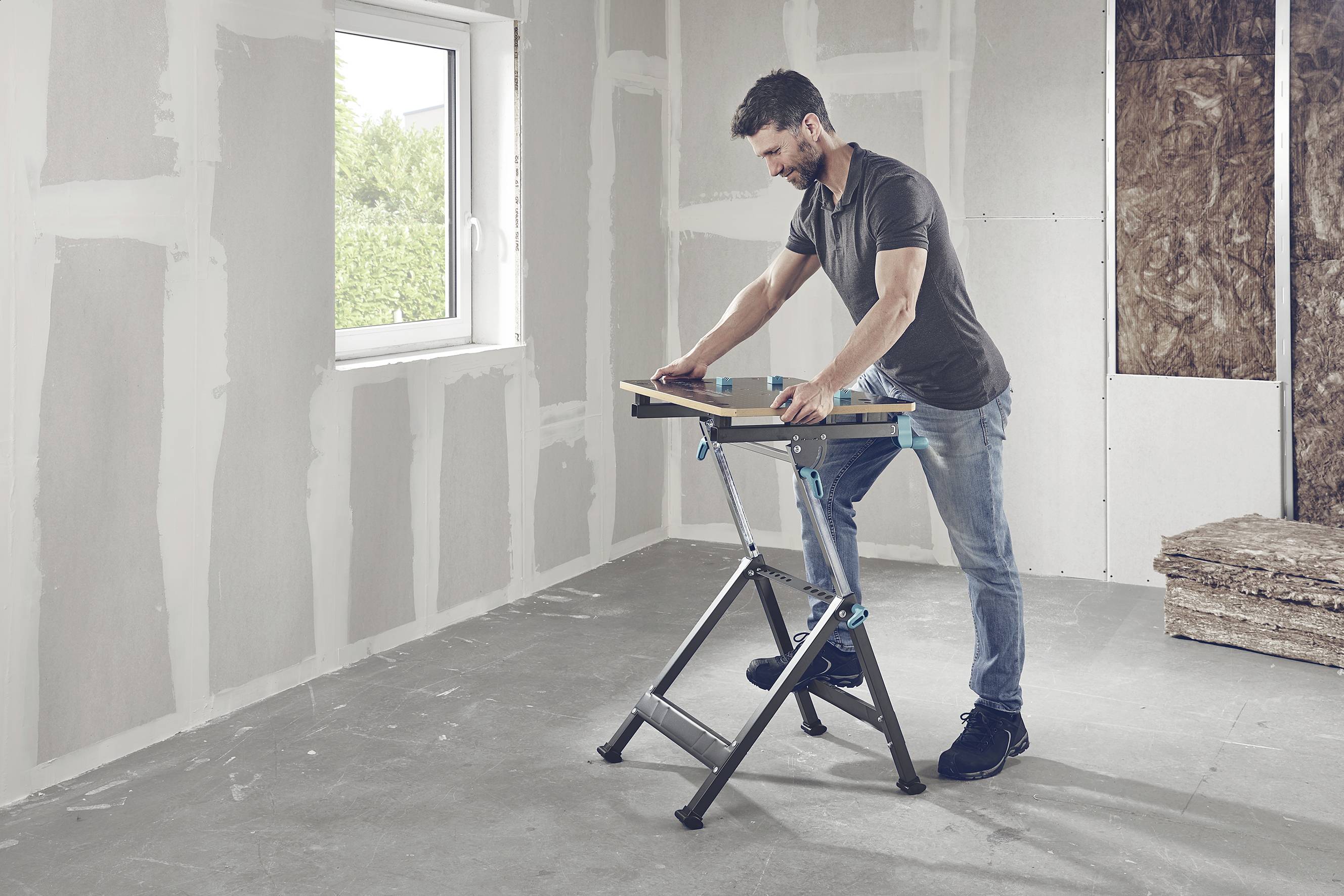 Ein Mann baut in einem unfertigen Raum etwas auf einem Klapptisch zusammen. Die Wände haben noch keine Tapete. Fenster zeigt grünen Außenbereich.