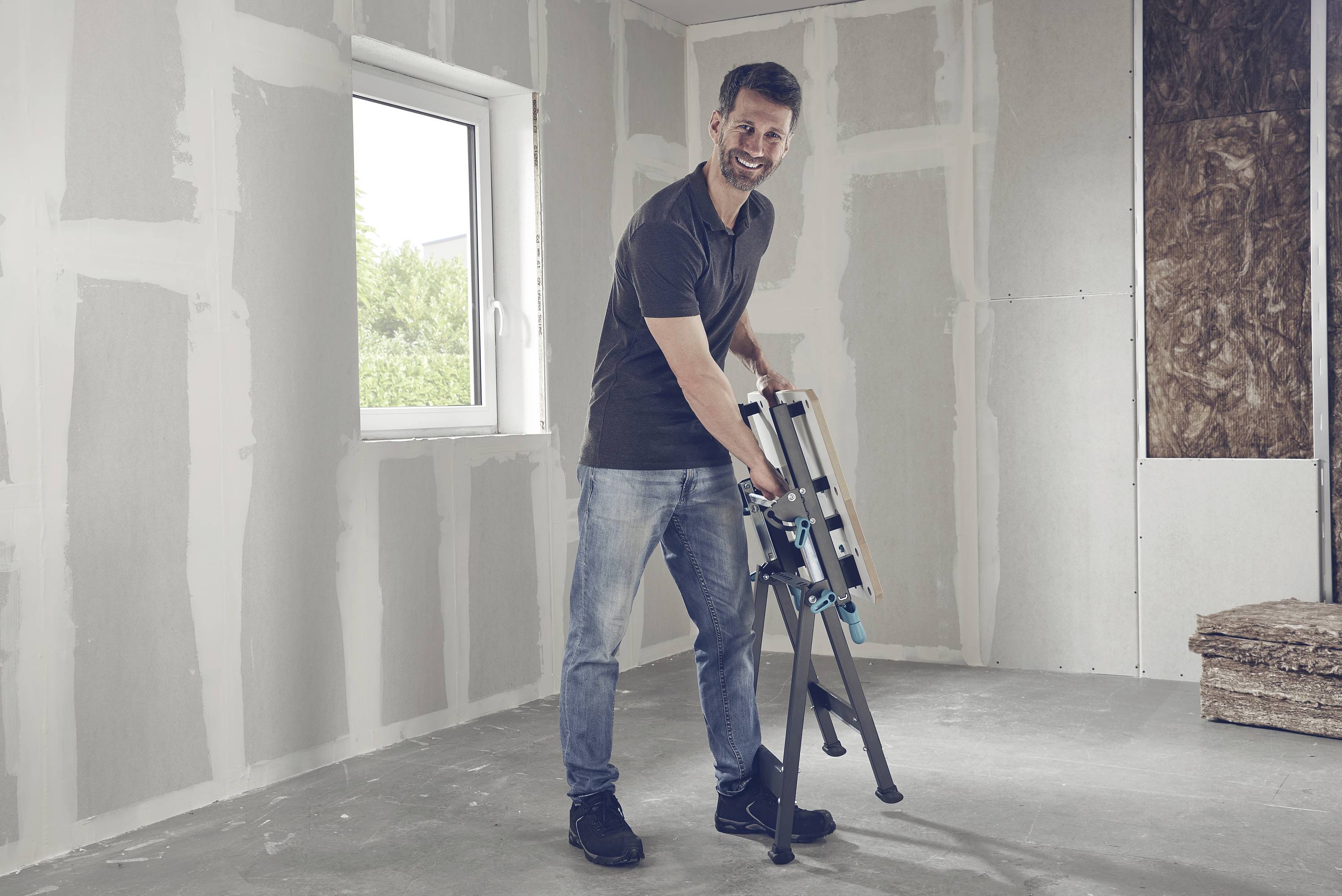 Ein Mann in Freizeitkleidung montiert eine Leiter in einem unfertigen Raum. Fenster mit Blick ins Freie, verputzte Wände im Hintergrund.