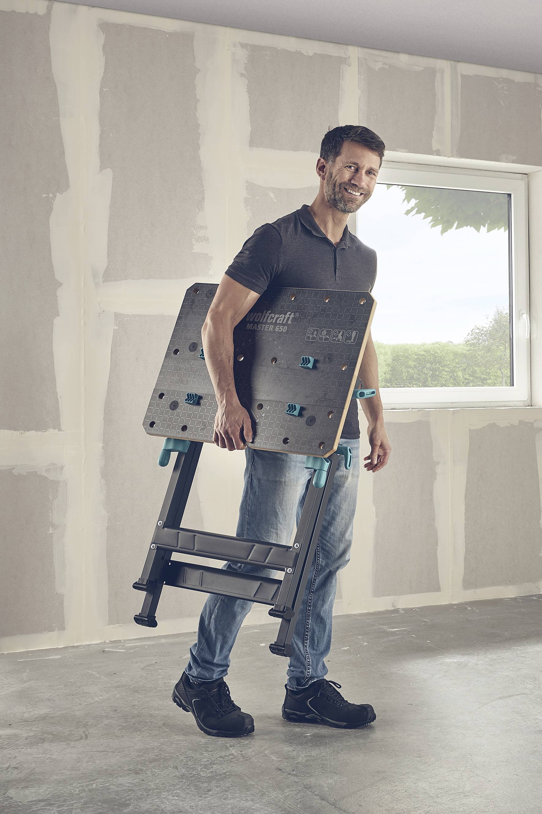 Ein Mann trägt eine zusammengeklappte Werkbank in einem renovierten Raum. Die Wände sind teils verputzt, Fenster im Hintergrund.