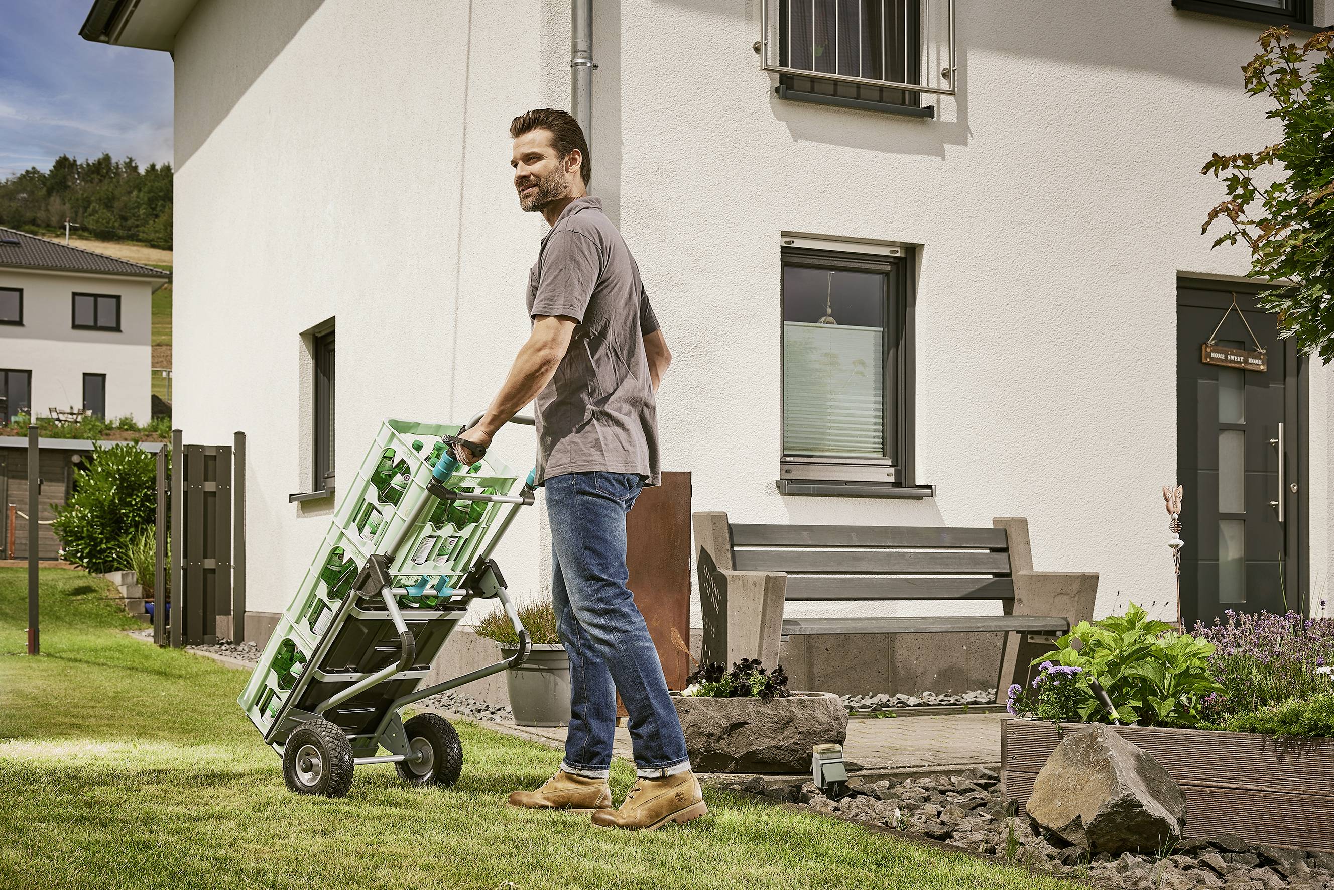 Ein Mann transportiert Getränkekisten auf einem Rollwagen über einen Rasen vor einem modernen Wohnhaus.