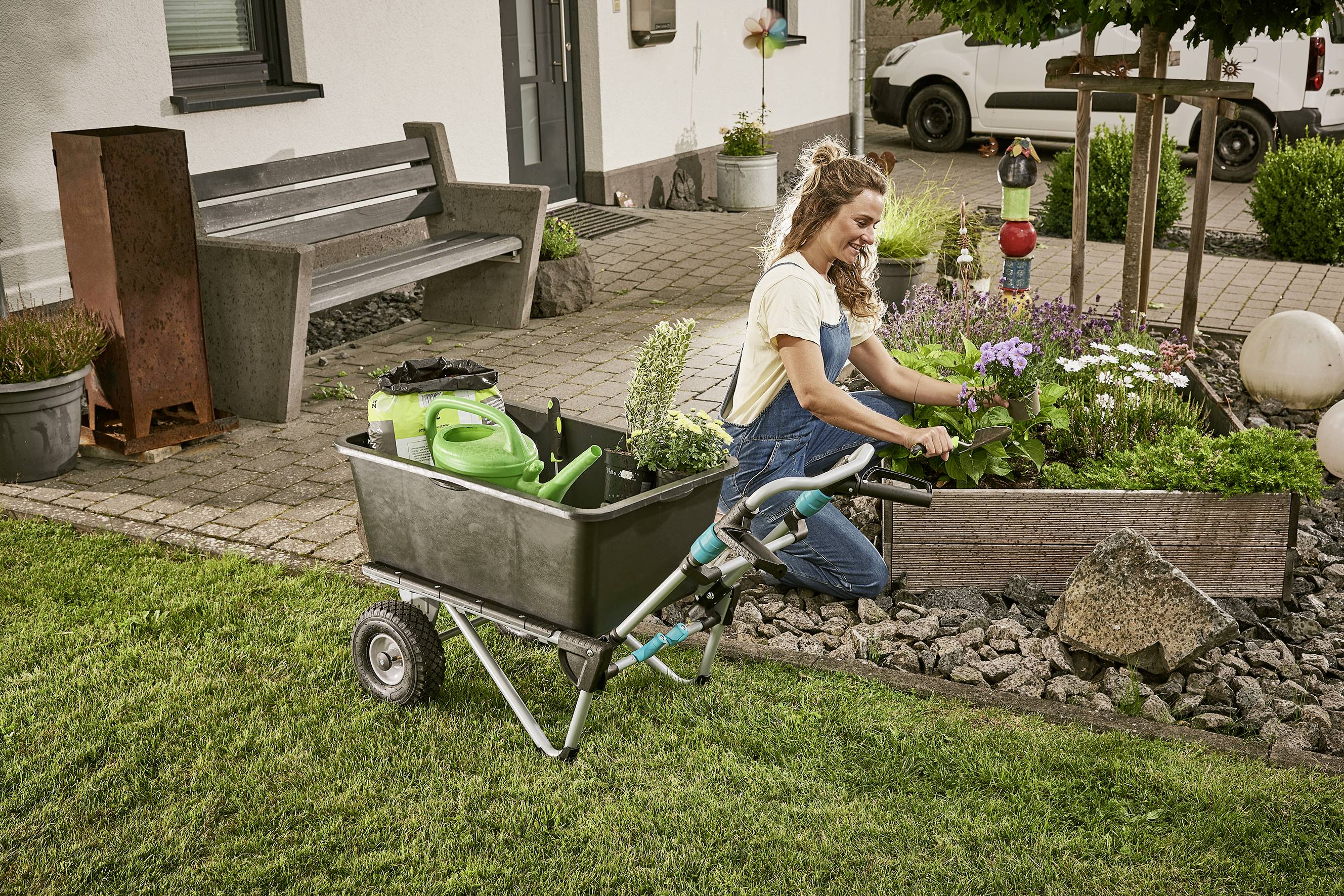 Eine Frau kniet in einem Garten und pflanzt Blumen in einem Beet. Neben ihr steht ein Gartensackkarren mit Gartengeräten.